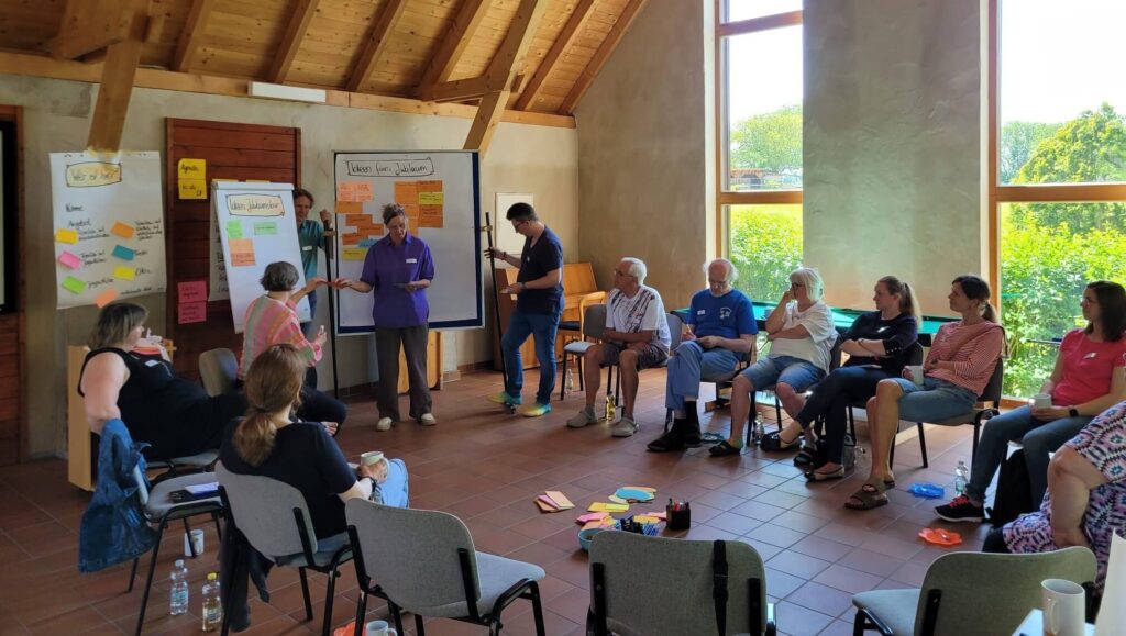 Teilnehmende des Aktiventreffs der DGhK OWL sitzen und stehen in einem Seminarraum auf dem Kirchberghof in Warburg und diskutieren gemeinsam an Stellwänden.